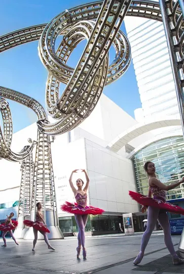 Ballet dancers in red tutus perform under a looping roller coaster structure in bright daylight.