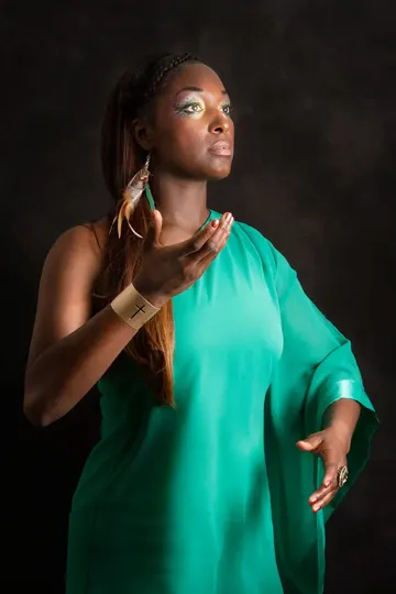Subject in teal dress, gold cross bracelet, feather earring, dark studio, soft lighting.