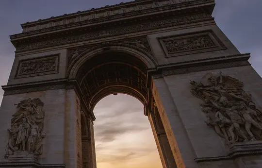 Arc de Triomphe at dusk, warm light illuminates the arch against a cloudy sky.