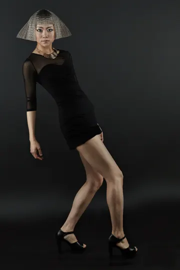 Subject in black dress and high heels, poised against dark backdrop with soft, directional lighting.