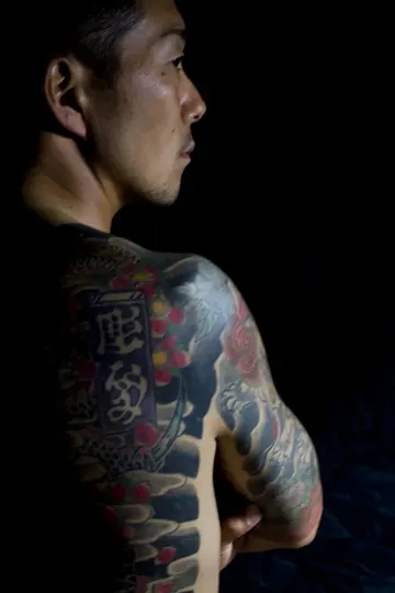 A man with intricate, colorful tattoos on his upper back and shoulder, profiled against a dark background with focused lighting.