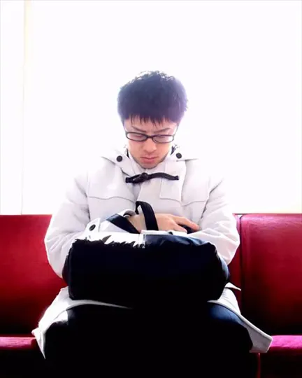 Subject in white coat sits on red couch, hands clasped, light streaming in.