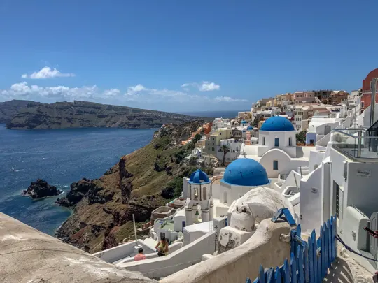 White-washed buildings with blue domes cascade down a cliffside, overlooking the Aegean Sea.