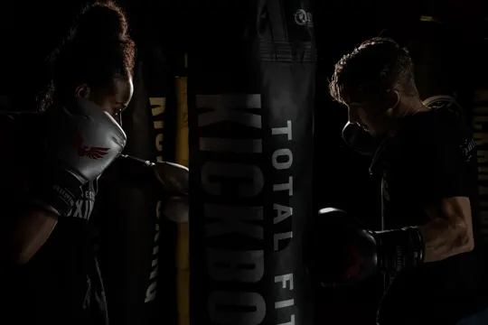 Two boxers in dim light, shadowed by a punching bag labeled "TOTAL FIT KICKBOXING.