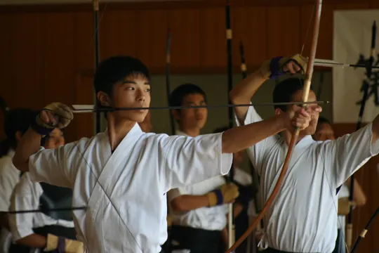 Archers in white uniforms draw bows in a wooden-floored dojo with soft, focused lighting.