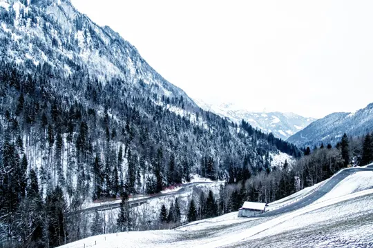 Snow-capped mountains and evergreen forests frame a winding road through a quiet, wintry valley.