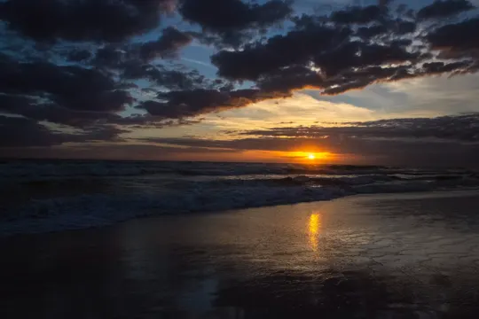 Sunset casts golden light on wet sand, waves, and dark clouds at the shore.