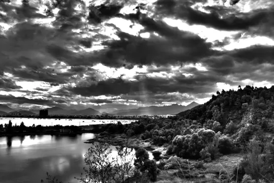 Dramatic clouds cast shifting shadows over a calm river and distant mountains.
