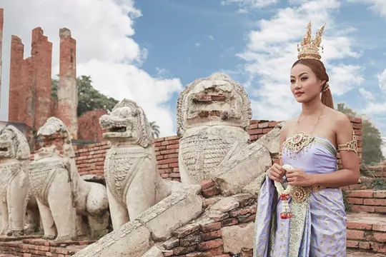 A woman in traditional attire stands before ancient stone lion statues and brick ruins under a bright sky.