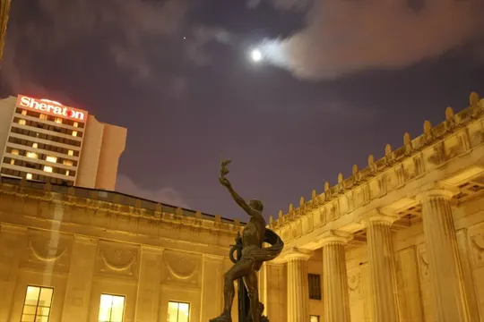 Bronze statue under a moonlit sky, with a Sheraton hotel and classical columns in the background.