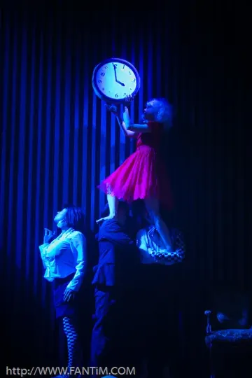 A woman in a red dress balances on a man's shoulders, reaching for a clock, illuminated by blue stage lighting.
