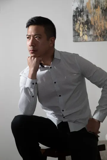 Subject in white shirt and black pants, seated, hand to chin, abstract painting in background.