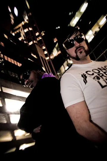 Two individuals, one in a white shirt with partial text visible, stand in a dimly lit urban environment at night, illuminated by artificial.
