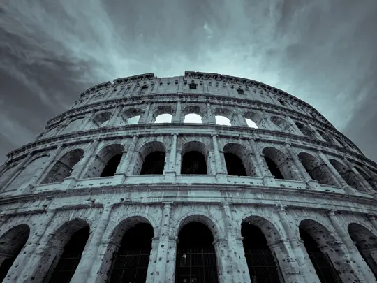 Ancient stone amphitheater rises under brooding sky, weathered arches framed by storm clouds.