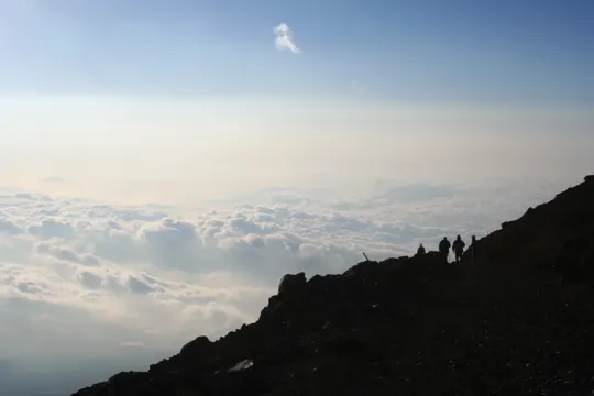 Silhouetted hikers traverse a rocky ridge above a sea of clouds under a clear sky.