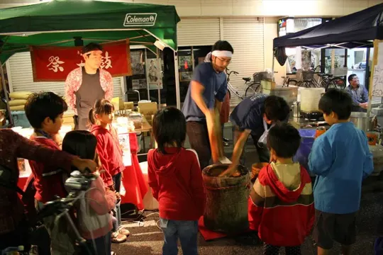Street vendor prepares food under green tent, children observe, warm light.