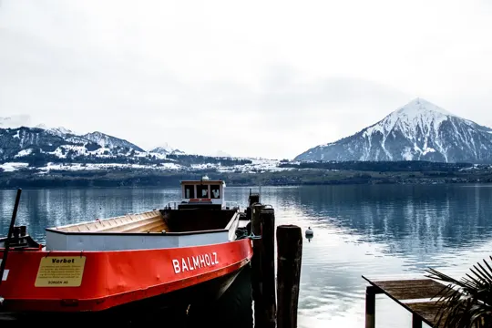 A red boat named "BALMHOLZ" rests at a wooden dock, framed by snow-capped mountains and a calm lake under an overcast sky.