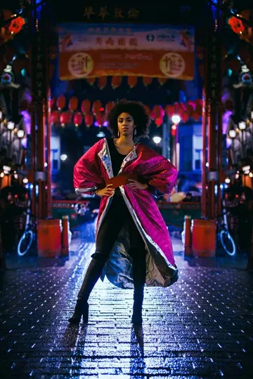 A figure in a magenta coat stands on a wet, cobblestone street under vibrant, traditional lanterns.