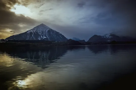 Sunlight pierces storm clouds, illuminating a snow-capped mountain reflected in still waters.