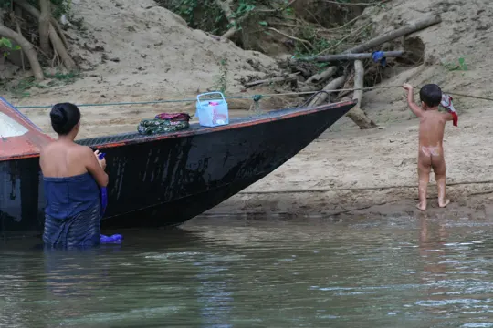 A child and adult near a boat on a sandy riverbank, soft light.