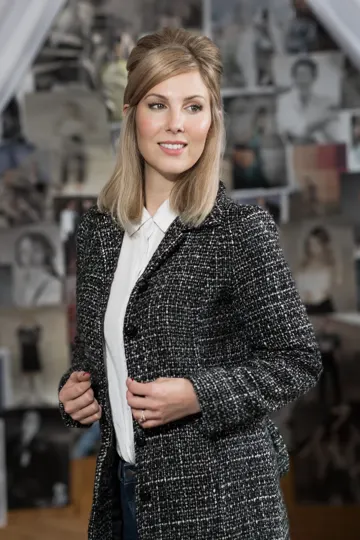 Blonde woman in tweed jacket stands before a backdrop of fashion sketches.