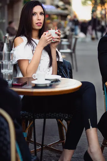 Woman in white cold-shoulder top sips coffee at outdoor café, soft natural light.