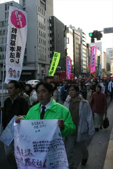 Protesters march through a city street, banners and signs visible, daylight.