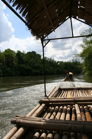 A bamboo raft glides along a river, framed by a thatched roof and distant greenery under a partly cloudy sky.