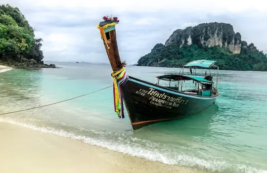 A traditional long-tail boat rests on a sandy beach, framed by turquoise waters and misty, forested limestone cliffs under an overcast sky.