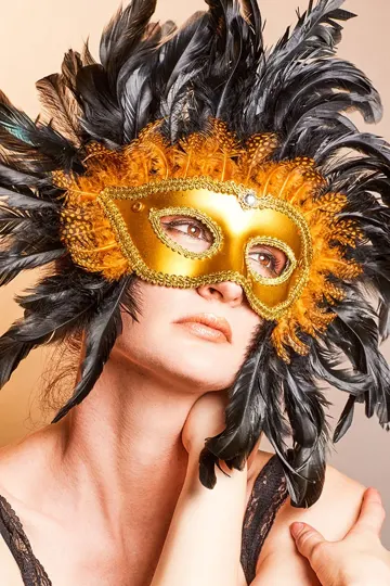 Subject wears ornate gold mask and black feather headdress, soft studio lighting.