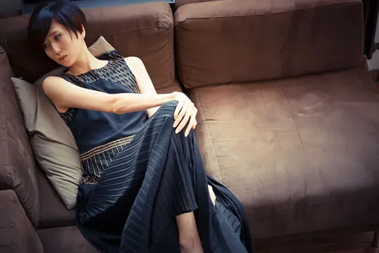 A woman in a patterned dress reclines on a brown sofa, softly lit.