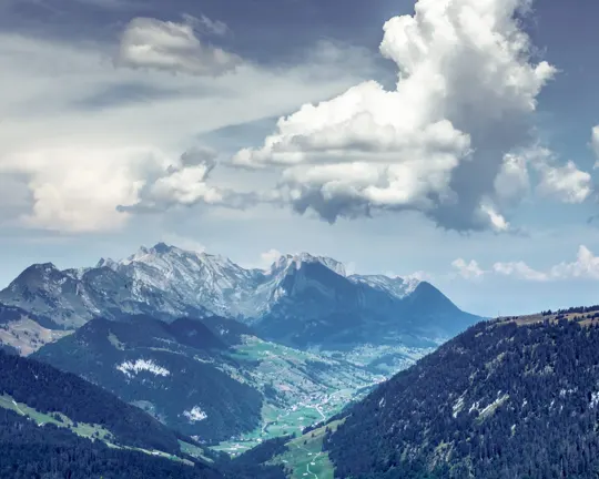 Sunlit mountain peaks rise under a dramatic sky, with valleys and forests below.